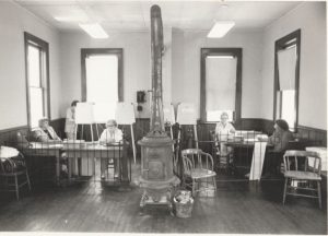 Historic black-and-white photo of a room with a central cast-iron stove and people seated at tables along the walls.